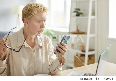 Focused woman with blond hair looking at smartphone screen while sitting at desk on workplace. Focused woman with blond hair looking at smartphone screen while sitting at desk on workplace. 134023694