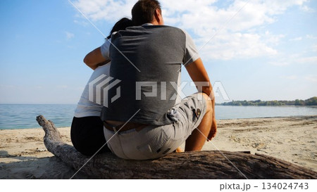 Happy pair hugs during sitting on wooden log on beach. Romantic couple admiring sea landscape at sunny day. Lovers spending time together at resort. Concept of summer vacation or holiday. Slow mo Happy pair hugs during sitting on wooden log on beach. Romantic couple admiring sea landscape at sunny day. Lovers spending time together at resort. Concept of summer vacation or holiday. Slow mo 134024743