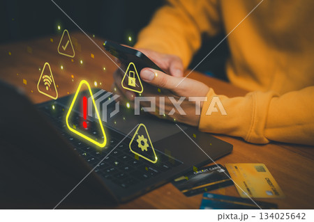 Person using smartphone and laptop with warning signs and cybersecurity icons, symbolizing online fraud, data breach, identity theft, internet security risks, and digital safety. 134025642