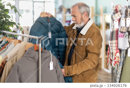 elderly man carefully chooses a down jacket in a store elderly man carefully chooses a down jacket in a store 134026176