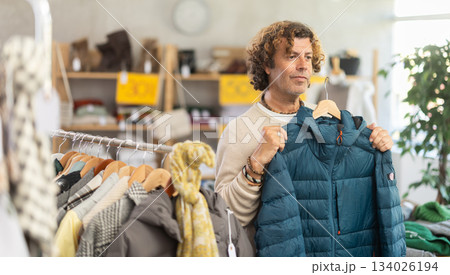 Stylish man standing in clothing shop and looking new down jacket made of high-quality material. Shopping and seasonal sales concept, winter season Stylish man standing in clothing shop and looking new down jacket made of high-quality material. Shopping and seasonal sales concept, winter season 134026194