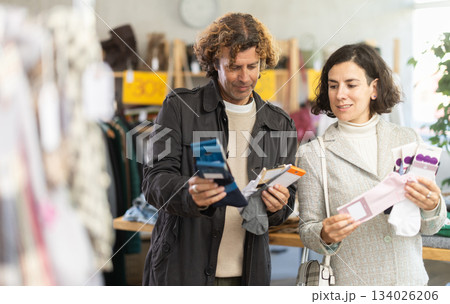 Positive couple shoppers examining and buying new warm socks in clothing store Positive couple shoppers examining and buying new warm socks in clothing store 134026206