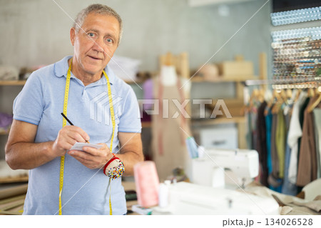 Senior man tailor in studio and writes down thoughts in notebook Senior man tailor in studio and writes down thoughts in notebook 134026528