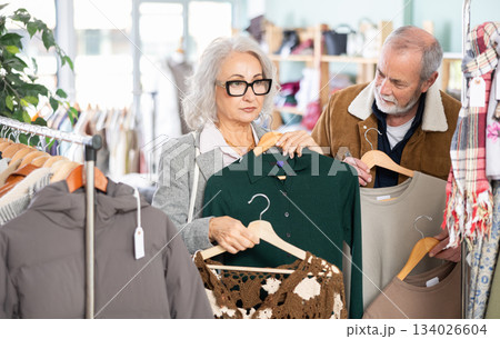Elderly married couple chooses fashionable warm dress in clothing store. Husband helps his wife choose dress Elderly married couple chooses fashionable warm dress in clothing store. Husband helps his wife choose dress 134026604