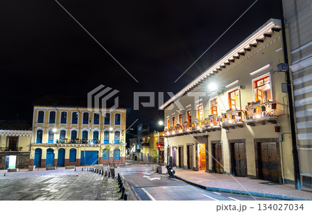 Colonial architecture lines San Francisco Square in historic center of Quito, Ecuador. White building features balconies with flower pots illuminated by streetlights under night sky 134027034