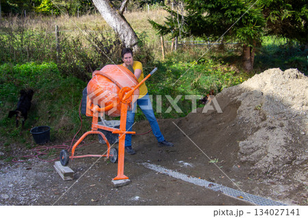 Woman puts cement with a shovel into the cement mixer. Woman puts cement with a shovel into the cement mixer. 134027141