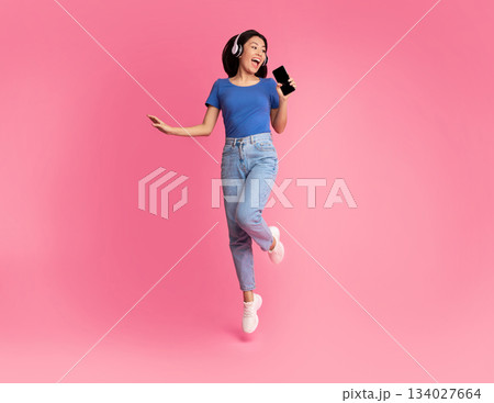 Carefree Singer. Asian lady singing favorite song and using cell phone as a mic, dancing and jumping, wearing wireless headphones. Excited woman having fun at pink studio, enjoying sound, full length Carefree Singer. Asian lady singing favorite song and using cell phone as a mic, dancing and jumping, wearing wireless headphones. Excited woman having fun at pink studio, enjoying sound, full length 134027664