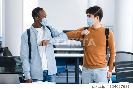 Elbow Bump. Smiling multicultural students wearing medical face masks greeting each other at classroom. Asian and african american men avoid touching, studying in university, standing at lecture hall Elbow Bump. Smiling multicultural students wearing medical face masks greeting each other at classroom. Asian and african american men avoid touching, studying in university, standing at lecture hall 134027681