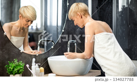 Woman Washing Face With Cold Water Standing Wrapped In Towel In Bathroom At Home. Selective Focus Woman Washing Face With Cold Water Standing Wrapped In Towel In Bathroom At Home. Selective Focus 134027788