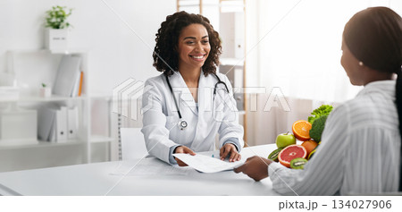 Personal diet. Cheerful african american female dietologist giving diet plan to woman client, clinic interior 134027906