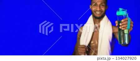 Workout Hydration. Black Athlete Man Offering Bottle Of Water To Camera Posing On Blue Studio Background. Shallow Depth, Selective Focus 134027920