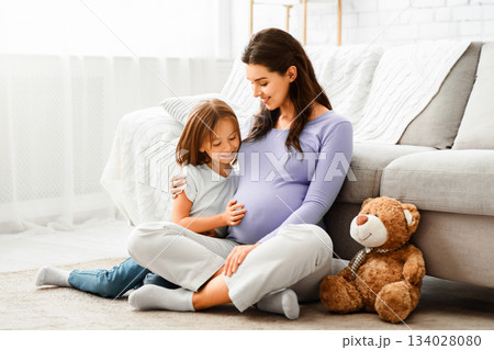 Family concept. Adorable little girl cuddling with her expecting mother at home, sitting on floor and playing with teddy bear, copy space 134028080