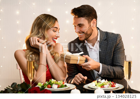 Happy man giving valentine's day present to his surprised girlfriend at romantic dinner in restaurant, pleased woman receiving gift box from boyfriend Happy man giving valentine's day present to his surprised girlfriend at romantic dinner in restaurant, pleased woman receiving gift box from boyfriend 134028125