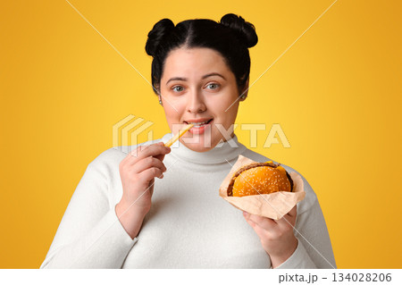 Causes Of Obesity. Young Overweight Woman Enjoying Fast Food, Eating Hamburger And French Fries, Posing Over Yellow Background With Empty Space 134028206