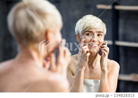 Happy Middle-Aged Woman Applying Facial Cream Moisturizer On Face Standing Wrapped In Towel In Bathroom. Mature Skin Treatment. Selective Focus Happy Middle-Aged Woman Applying Facial Cream Moisturizer On Face Standing Wrapped In Towel In Bathroom. Mature Skin Treatment. Selective Focus 134028313