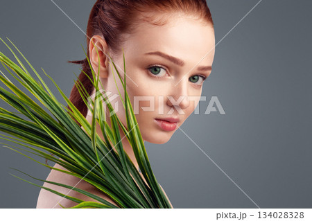 Sensual Ginger-Haired Young Woman Posing With Green Plant Standing In Studio On White Background, Looking At Camera. Female Portrait, Natural Beauty Of Youth Concept Sensual Ginger-Haired Young Woman Posing With Green Plant Standing In Studio On White Background, Looking At Camera. Female Portrait, Natural Beauty Of Youth Concept 134028328
