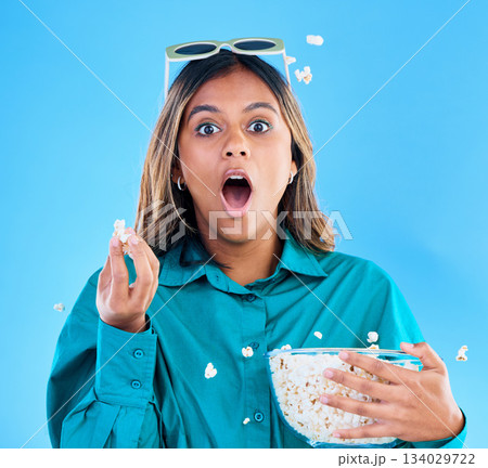 3D glasses, popcorn and wow with a woman in studio on a blue background looking shocked while eating a snack. Portrait, movie and video entertainment with an attractive young female feeling surprised 3D glasses, popcorn and wow with a woman in studio on a blue background looking shocked while eating a snack. Portrait, movie and video entertainment with an attractive young female feeling surprised 134029722