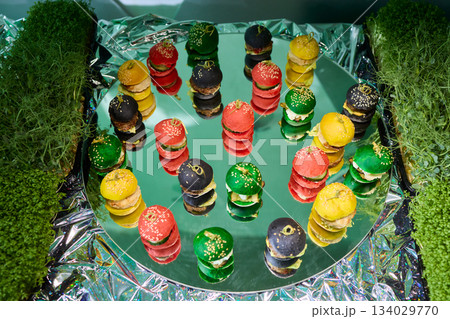 Colorful miniature burger macarons displayed on shiny green foil surface, Playfully arranged rainbow hued miniature burger macarons with glossy shells and festive appeal 134029770