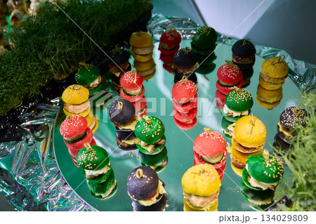 Colorful miniature burger macarons displayed on shiny green foil surface, Playfully arranged rainbow hued miniature burger macarons with glossy shells and festive appeal Colorful miniature burger macarons displayed on shiny green foil surface, Playfully arranged rainbow hued miniature burger macarons with glossy shells and festive appeal 134029809