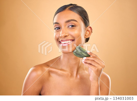 Face portrait, skincare and woman with gua sha in studio isolated on a brown background. Dermatology, facial massage or happy Indian female model with jade crystal or stone for healthy skin treatment 134031003