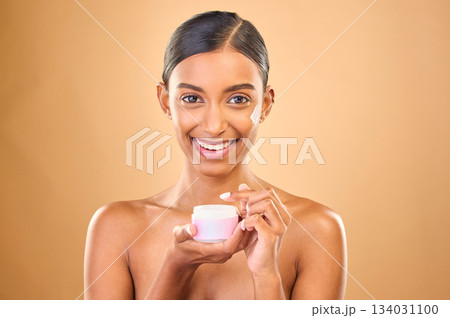 Face, skincare and woman with cream jar in studio isolated on a brown background. Dermatology cosmetics, portrait and happy Indian female apply lotion, creme and moisturizer product for healthy skin. 134031100