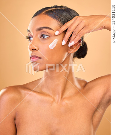 Face, skincare idea and woman with cream in studio isolated on a brown background. Dermatology, cosmetics and thinking Indian female model with lotion, beauty creme or moisturizer for skin health. 134031119