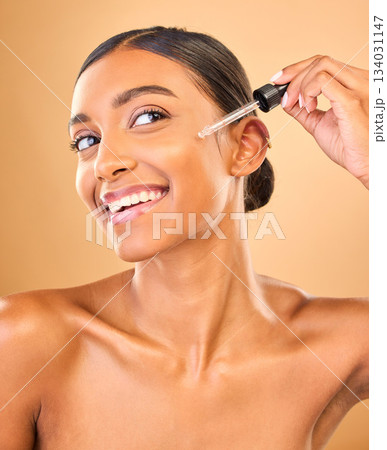 Face, skincare serum and happy woman in studio isolated on a brown background. Dermatology, cosmetics and smile of Indian female model with hyaluronic acid, retinol or essential oil for healthy skin. Face, skincare serum and happy woman in studio isolated on a brown background. Dermatology, cosmetics and smile of Indian female model with hyaluronic acid, retinol or essential oil for healthy skin. 134031147
