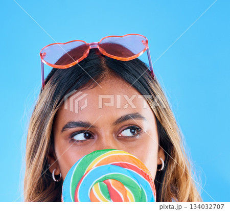 Thinking, lollipop and a woman on a blue background in studio wearing heart glasses for fashion. Idea, candy and sweet with an attractive young female eating a giant snack while feeling thoughtful Thinking, lollipop and a woman on a blue background in studio wearing heart glasses for fashion. Idea, candy and sweet with an attractive young female eating a giant snack while feeling thoughtful 134032707