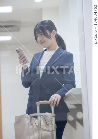 ガラス越しの窓にスマホを見るビジネス女性の立ち姿・出張移動と情報確認シーンのライフスタイル 134034762