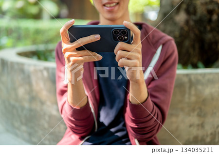 A man watching video or playing game on smartphone holding horizontally with both hands in the park. A man watching video or playing game on smartphone holding horizontally with both hands in the park. 134035211