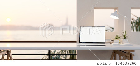 White screen laptop and a coffee cup on white table under lamp light near a terrace with river view. 134035260