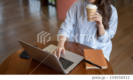 Woman in blue shirt typing on laptop while holding coffee cup and sitting at wooden table in cafe. Woman in blue shirt typing on laptop while holding coffee cup and sitting at wooden table in cafe. 134038412