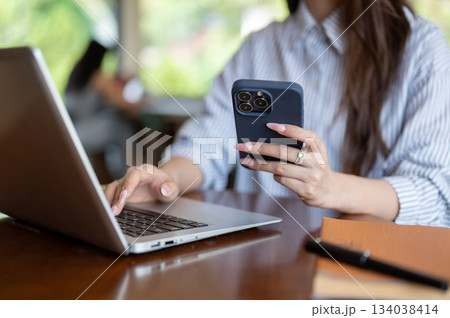 Close up of hands with long nails holding a phone and typing on laptop while sitting at wooden table Close up of hands with long nails holding a phone and typing on laptop while sitting at wooden table 134038414