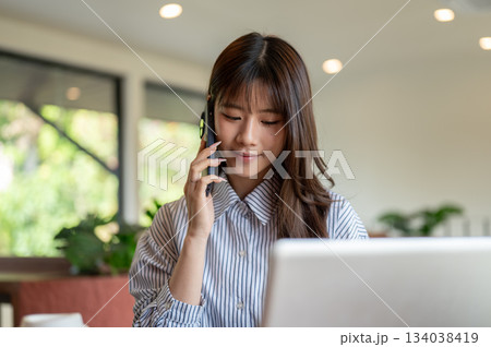 Asian woman in blue striped shirt talking on phone while sitting in front of laptop at table in cafe Asian woman in blue striped shirt talking on phone while sitting in front of laptop at table in cafe 134038419