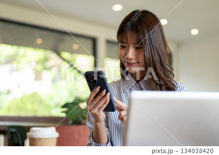 Blue striped shirt asian woman looking and typing on phone while sitting in front of laptop in cafe. Blue striped shirt asian woman looking and typing on phone while sitting in front of laptop in cafe. 134038420