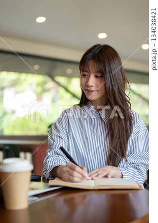 Asian woman writing in notebook with fountain pen and looking at laptop while sitting in the cafe. Asian woman writing in notebook with fountain pen and looking at laptop while sitting in the cafe. 134038421