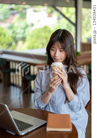 Smiling asian woman in blue shirt looking at laptop while holding coffee sitting at cafe wood table Smiling asian woman in blue shirt looking at laptop while holding coffee sitting at cafe wood table 134038431