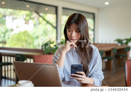 Asian woman with a hand under chin holding phone and looking at laptop while sitting at wooden table Asian woman with a hand under chin holding phone and looking at laptop while sitting at wooden table 134038434