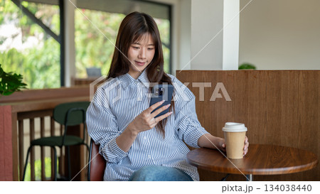 Smiling asian woman looking at phone and holding coffee while sitting at wooden stool table in cafe. 134038440