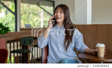 Smiling asian woman talking on the phone and grabbing coffee cup while sitting at cafe wooden stool Smiling asian woman talking on the phone and grabbing coffee cup while sitting at cafe wooden stool 134038442