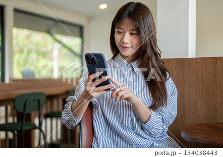 Smiling asian woman in blue shirt looking and typing on her phone while sitting at cafe wooden table Smiling asian woman in blue shirt looking and typing on her phone while sitting at cafe wooden table 134038443
