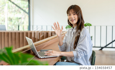 Blue shirt asian woman smiling and waving a hand while sitting at wooden counter in cafe or library 134038457
