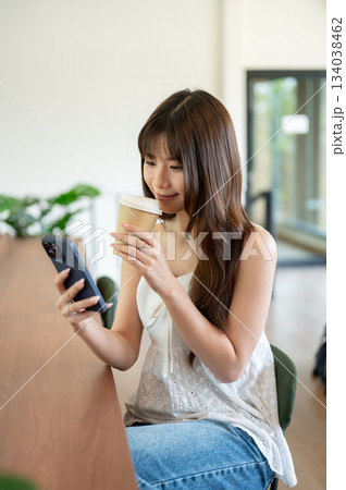 Asian woman in camisole sipping coffee and looking at her phone while sitting at cafe wooden counter 134038462