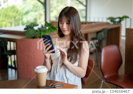 Asian woman in camisole smiling and looking at phone in hands while sitting at cafe's wooden table. Asian woman in camisole smiling and looking at phone in hands while sitting at cafe's wooden table. 134038467