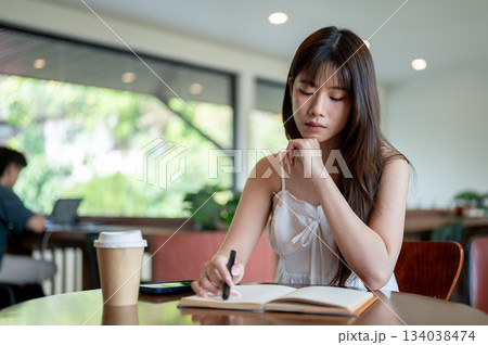 Asian woman in camisole holding pen and reading book while sitting with phone and coffee on table. Asian woman in camisole holding pen and reading book while sitting with phone and coffee on table. 134038474