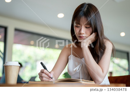Asian woman put a hand under chin holding pen writing in notebook while sitting with coffee on table Asian woman put a hand under chin holding pen writing in notebook while sitting with coffee on table 134038478