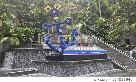 A vibrant anchor sculpture and cruise ship model sit amid cascading water and lush greenery, blending nautical art with tropical serenity in a visually captivating indoor garden sett 134038940