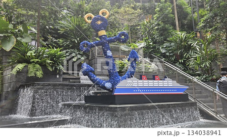 A vibrant anchor sculpture and cruise ship model sit amid cascading water and lush greenery, blending nautical art with tropical serenity in a visually captivating indoor garden sett 134038941