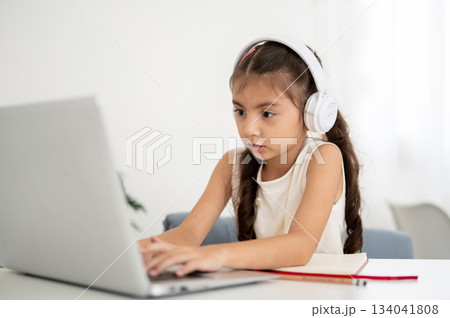 Asian girl kid wearing headphones looking and typing on laptop while sitting at white table in house 134041808