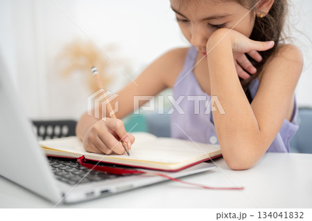 Close up of asian kid girl writing in notebook over a laptop while sitting at white table in house. Close up of asian kid girl writing in notebook over a laptop while sitting at white table in house. 134041832